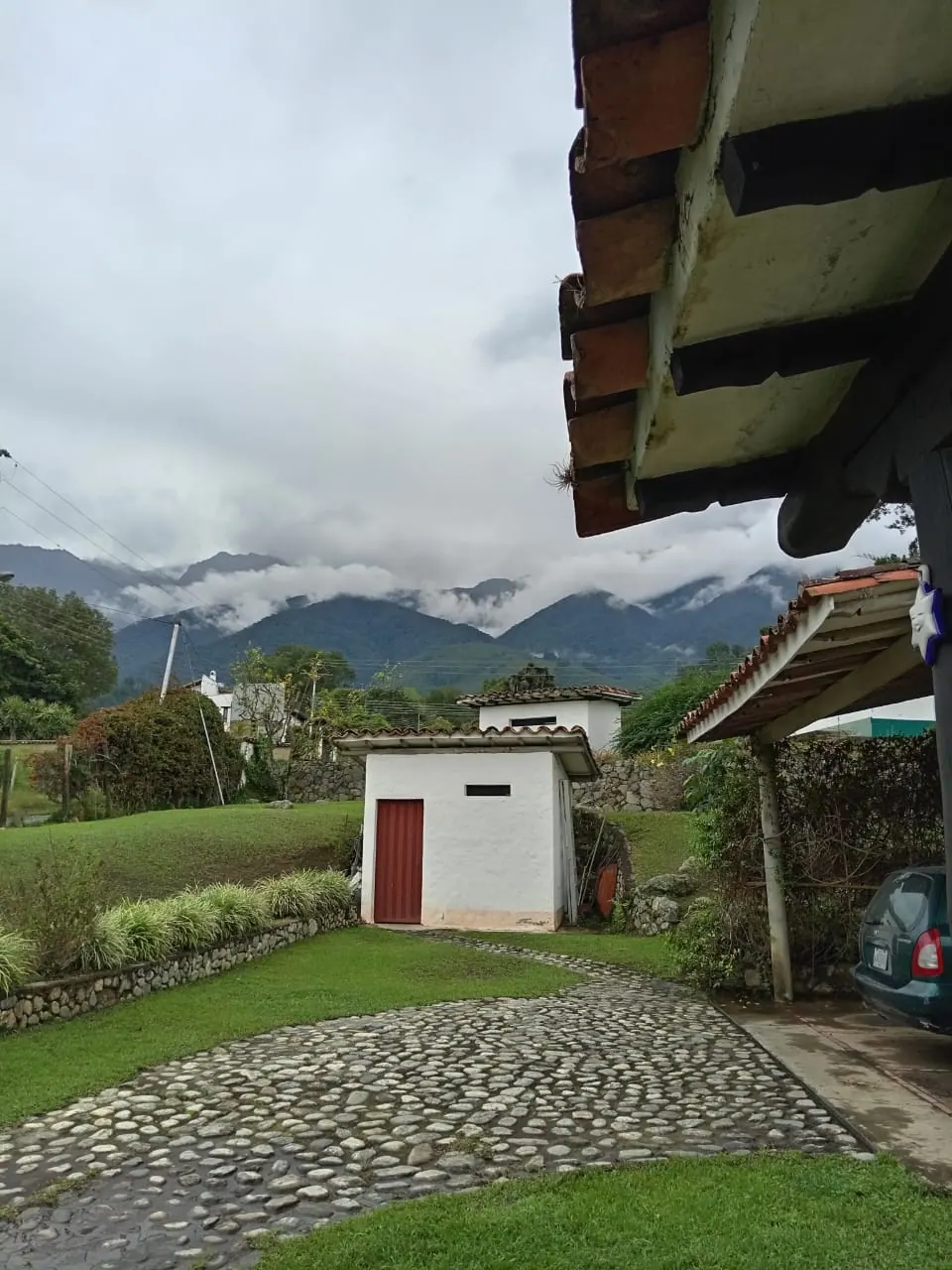 Entrada empedrada con casa blanca, jardín y montañas nubladas al fondo